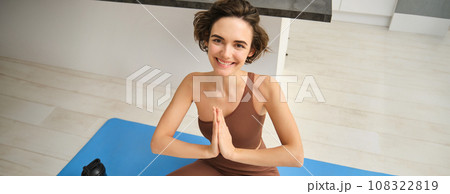 Young healthy beautiful woman in sports top and leggings practicing yoga at home, sitting in lotus position on yoga mat, meditating, smiling, relaxed, eyes closed, meditation concept 108322819