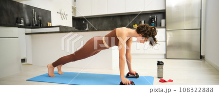 Woman workout from home, stands in plunk on Push Up Bars Stand, grips handle for chest press, doing gym exercises on kitchen floor Woman workout from home, stands in plunk on Push Up Bars Stand, grips handle for chest press, doing gym exercises on kitchen floor 108322888
