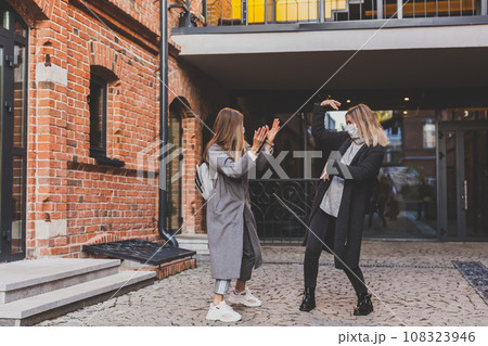 Two young stylish women in medical masks dancing and having fun while walking the street in town. Friends strolling outdoor in city Two young stylish women in medical masks dancing and having fun while walking the street in town. Friends strolling outdoor in city 108323946