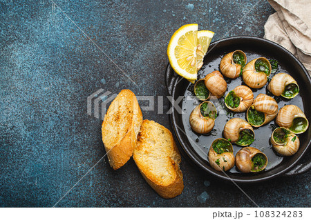 Escargots de Bourgogne Snails with Garlic Butter and Parsley in black cast iron pan with Lemon and Toasted Baguette Slices on rustic stone background top view, traditional French Delicacy, copy space. Escargots de Bourgogne Snails with Garlic Butter and Parsley in black cast iron pan with Lemon and Toasted Baguette Slices on rustic stone background top view, traditional French Delicacy, copy space. 108324283