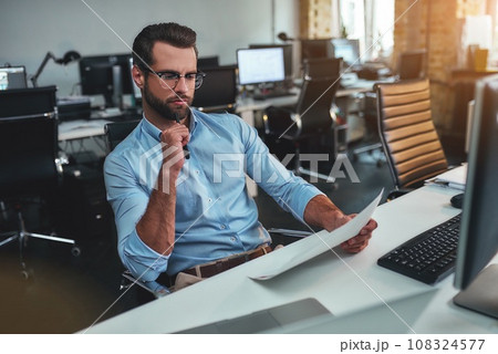 New strategy. Concentrated bearded man in eyeglasses and formal wear looking at documents and thinking while sitting in the modern office 108324577