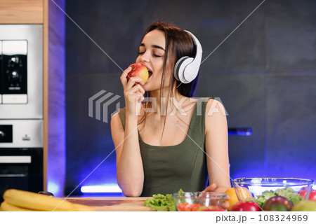 Young attractive woman eats red apple and listens to the music at kitchen in morning. Healthy lifestyle concept, enjoying breakfast. Young attractive woman eats red apple and listens to the music at kitchen in morning. Healthy lifestyle concept, enjoying breakfast. 108324969