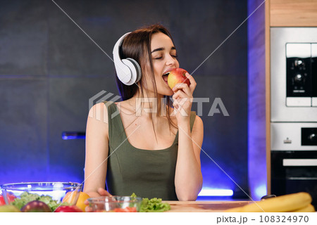 Young attractive woman eats red apple and listens to the music at kitchen in morning. Healthy lifestyle concept, enjoying breakfast. Young attractive woman eats red apple and listens to the music at kitchen in morning. Healthy lifestyle concept, enjoying breakfast. 108324970