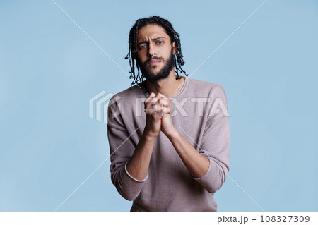 Arab man with clenched hands looking at camera with pleading facial expression. Person praying with folded arms gesture while asking for forgiveness with hopeful emotions Arab man with clenched hands looking at camera with pleading facial expression. Person praying with folded arms gesture while asking for forgiveness with hopeful emotions 108327309