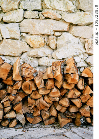 Stack of firewood is stacked against the stone wall of a house on a paved path 108328859