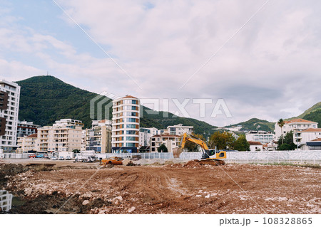 Yellow excavator digs the earth with a bucket at a construction site near high-rise buildings 108328865