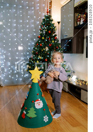 Little girl with a teddy bear stands near a toy Christmas tree in the room 108328910