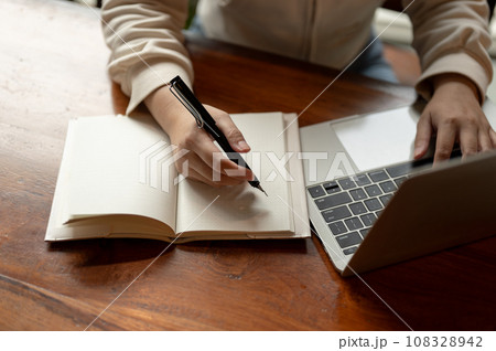Cropped image a woman working on her laptop and taking notes in her notebook at a desk. Cropped image a woman working on her laptop and taking notes in her notebook at a desk. 108328942