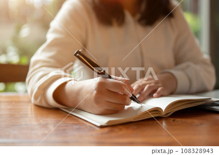 Cropped image of an Asian woman in casual clothes writing something in her book at a table indoors. 108328943