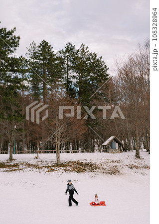 Mom is carrying a small child in a sled past a wooded hill with a stone shed. Side view Mom is carrying a small child in a sled past a wooded hill with a stone shed. Side view 108328964