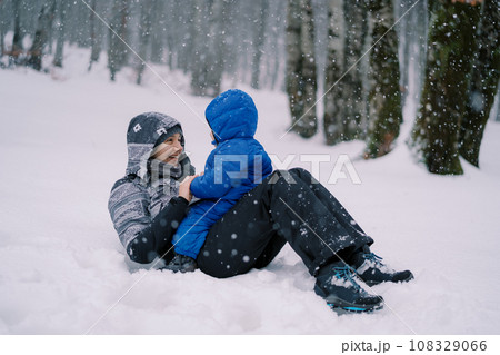 Laughing mom lies in the snow with a small child sitting on her belly under the snowfall 108329066