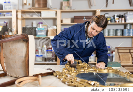 Skilled middle-aged male furniture repair specialist in uniform restoring Victorian mirror frame during workday in workshop Skilled middle-aged male furniture repair specialist in uniform restoring Victorian mirror frame during workday in workshop 108332695