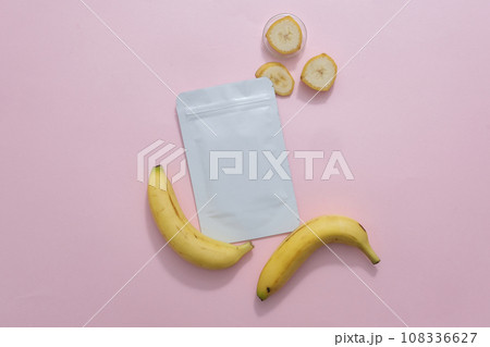 Top view of a facial sheet mask package decorated with banana and banana slices on a pink background. Minimalist concept for advertising cosmetic with natural ingredient Top view of a facial sheet mask package decorated with banana and banana slices on a pink background. Minimalist concept for advertising cosmetic with natural ingredient 108336627