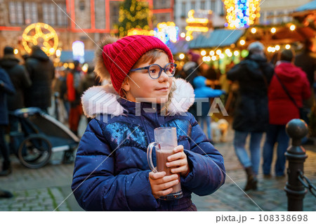 Little cute preschool girl drinking hot children punch or chocolate on German Christmas market. Happy child on traditional family market in Germany, Laughing kid in colorful winter clothes 108338698