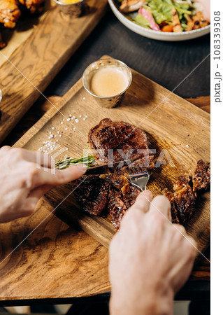 Close up of hands holding knife and fork is cutting grilled ribeye steak on wooden cutting board Close up of hands holding knife and fork is cutting grilled ribeye steak on wooden cutting board 108339308
