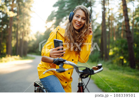 Happy woman with headphones, smartphone rides bicycle in sunny park. Active lifestyle, resting. Happy woman with headphones, smartphone rides bicycle in sunny park. Active lifestyle, resting. 108339501
