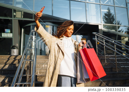 Young woman standing along road holding shopping bags, using mobile phone. Discounts, sale concept. Young woman standing along road holding shopping bags, using mobile phone. Discounts, sale concept. 108339835