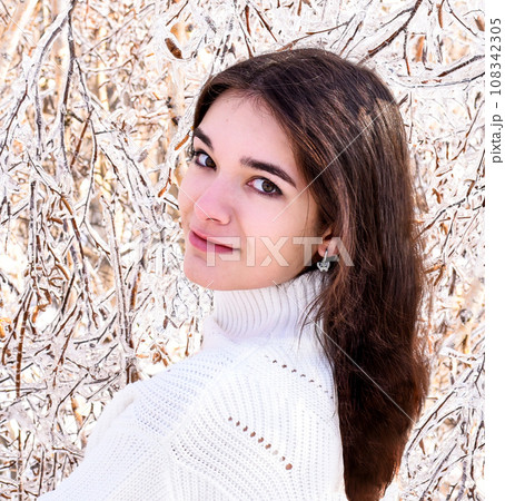 Portrait of young Russian woman in white sweater looking at camera next to frozen branches of birch trees, covered with ice. Winter forest in Russia Portrait of young Russian woman in white sweater looking at camera next to frozen branches of birch trees, covered with ice. Winter forest in Russia 108342305