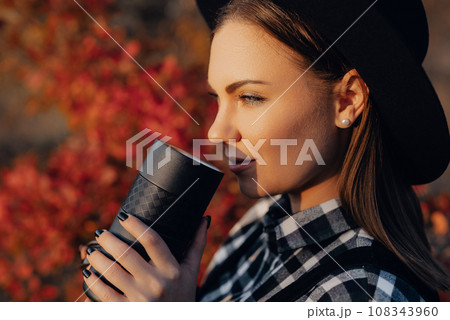 Hipster woman enjoys aroma fresh coffee. Drinking from thermos in autumn forest 108343960