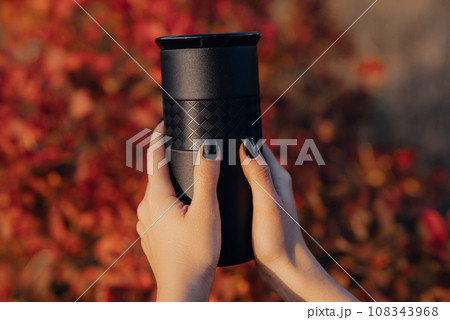 Woman hands holds black thermos cup with fresh coffee. Drinking in autumn forest 108343968