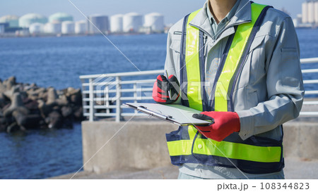 クリップボードを持つ作業服の男性・海と工業地帯│建設業・製造業・プラント　イメージ 108344823