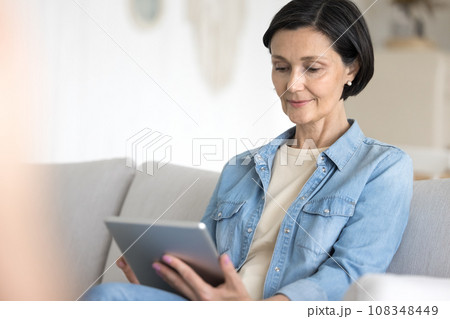 Mature woman working on tablet seated on cozy couch Mature woman working on tablet seated on cozy couch 108348449