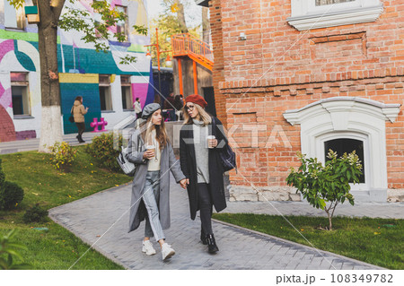 Two girls walking on street together and holding their hands. They are wearing spring or autumn clothes and they are happy. Friendship and relationship concept 108349782