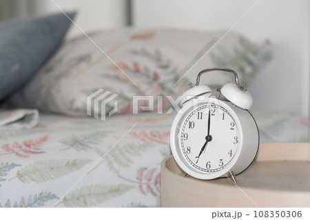 Close-up of a round white alarm clock on a table in the bedroom. The hands on the clock show seven o'clock in the morning, time to get up. Retro alarm clock on the table, vintage tone. space for text 108350306