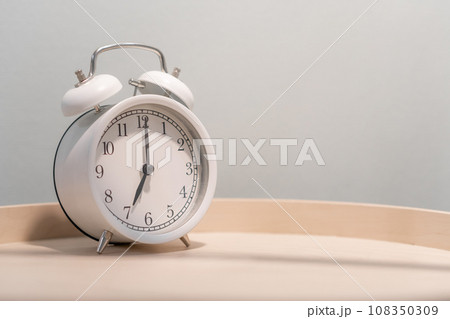 Close-up of a round white alarm clock on a table in the bedroom. The hands on the clock show seven o'clock in the morning, time to get up. Retro alarm clock on the table, vintage tone. space for text 108350309
