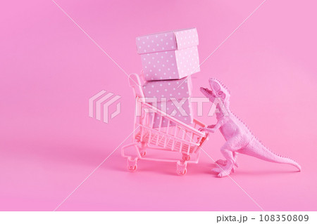 Pink Friday concept. Funny pink dinosaur toy with shopping cart full of present boxes on pink background 108350809