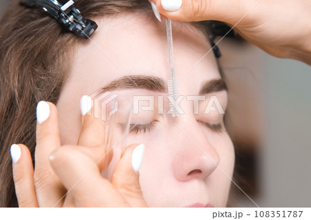 Painting eyebrows with an eyebrow brush. Eyebrow care, eyebrow lamination. close-up of the master's hands with a microbrush, which combs the model's eyebrows after the procedure Painting eyebrows with an eyebrow brush. Eyebrow care, eyebrow lamination. close-up of the master's hands with a microbrush, which combs the model's eyebrows after the procedure 108351787