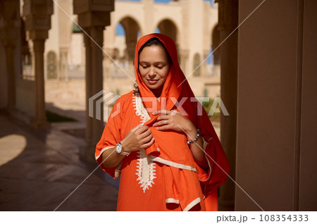 Beautiful woman in Moroccan traditional dress and head scarf, against beige marble columns and arches background 108354333