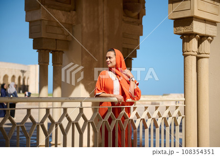 Gorgeous Middle Eastern Muslim woman with head covered in hijab and ethnic dress, smiles looking aside, Hassan II mosque 108354351