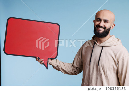 Smiling arab man holding speech bubble for product advertising mock up portrait. Cheerful young person posing with speak balloon with copy space mock up and looking at camera Smiling arab man holding speech bubble for product advertising mock up portrait. Cheerful young person posing with speak balloon with copy space mock up and looking at camera 108356188