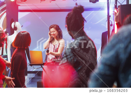 Smiling woman dj performing on stage while people dancing in nightclub at live concert. Young musician singing in microphone and mixing music at discotheque party in club Smiling woman dj performing on stage while people dancing in nightclub at live concert. Young musician singing in microphone and mixing music at discotheque party in club 108356203