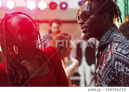 Smiling african american couple laughing and chatting in dark nightclub illuminated with vibrant lights. Carefree man and woman talking and socializing while partying in club 108356210