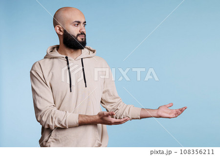 Arab man pointing to left while advertising product with neutral facial expression. Young person wearing casual hoodie clothes showing with hands and looking to side while posing in studio 108356211