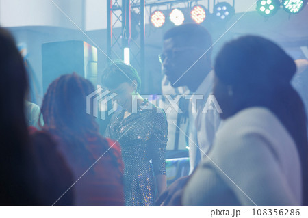 Smiling people crowd dancing and partying on dancefloor in nightclub. Carefree clubbers having fun, socializing and enjoying discotheque in club with colorful lights in fog 108356286