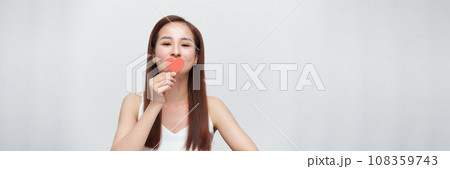 Woman in love, holding a little paper heart over eye, standing over white background, panorama 108359743