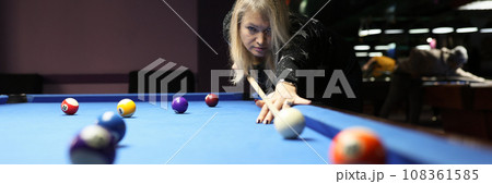 Focused woman hits white ball leaning above blue pool table Focused woman hits white ball leaning above blue pool table 108361585