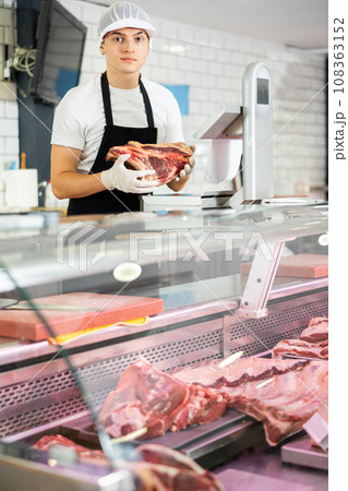 Positive young salesman demonstrating piece of meat in butcher shop Positive young salesman demonstrating piece of meat in butcher shop 108363152