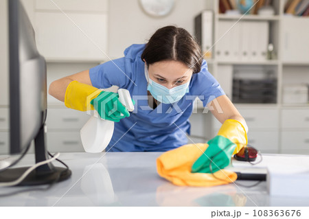 Woman in protective mask wipes dust from table in office Woman in protective mask wipes dust from table in office 108363676