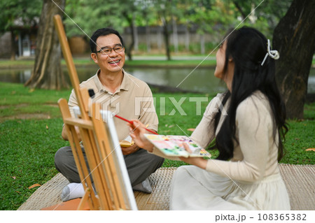 Happy 50s man and young adult daughter painting picture on the green grass at park 108365382
