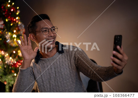 A cheerful Asian man is talking on a video call with his friend or family on a Christmas night. 108366477