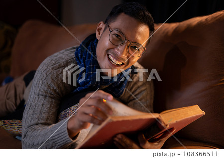 A happy Asian man in cosy clothes is reading a book while lying on sofa in his living room at night. 108366511
