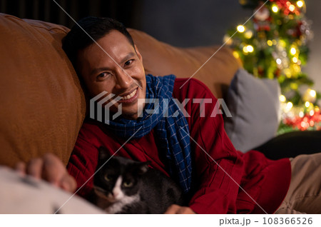 A happy Asian man is lying on a sofa with his cat, celebrating Christmas night at home together. A happy Asian man is lying on a sofa with his cat, celebrating Christmas night at home together. 108366526