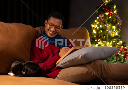 A relaxed and happy Asian man is admiring his sleeping cat while sitting on a couch together. 108366539