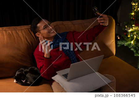 A happy Asian man enjoys talking on a video call on his phone with his friend while lying on a couch A happy Asian man enjoys talking on a video call on his phone with his friend while lying on a couch 108366545