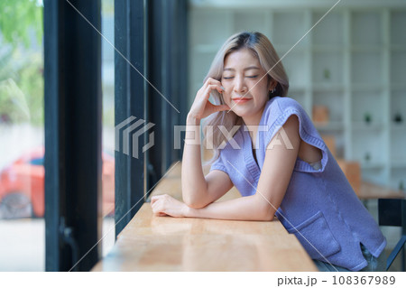 Asian young woman making notes in paper notebook sitting on cozy sofa on background of window. Pretty female writing notes in diary or making to do list relaxing at home 108367989