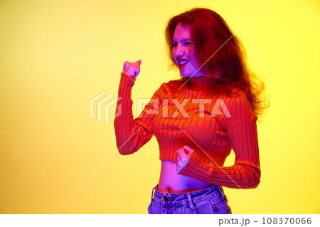 Young girl expressing excitement and cheering emotions against yellow studio background in neon light Young girl expressing excitement and cheering emotions against yellow studio background in neon light 108370066